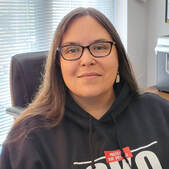 Jess McPherson, a medium complected woman with long dark brown hair, glasses, shell earrings, wears a black hoodie with the words 